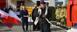 Przekazanie pojazdu ratowniczo-gaśniczego do OSP Śląskowo. Zdjęcie przedstawia plac przed remizą OSP w Śląskowie. Na tle budynku oraz uczestników uroczystości, kapłan dokonuje poświęcenia przekazanego pojazdu ratowniczo-gaśniczego. W prawej ręce trzyma kropidło. Z lewej strony znajduje się stojak z flagami, a z prawej widoczny jest fragment wozu strażackiego.