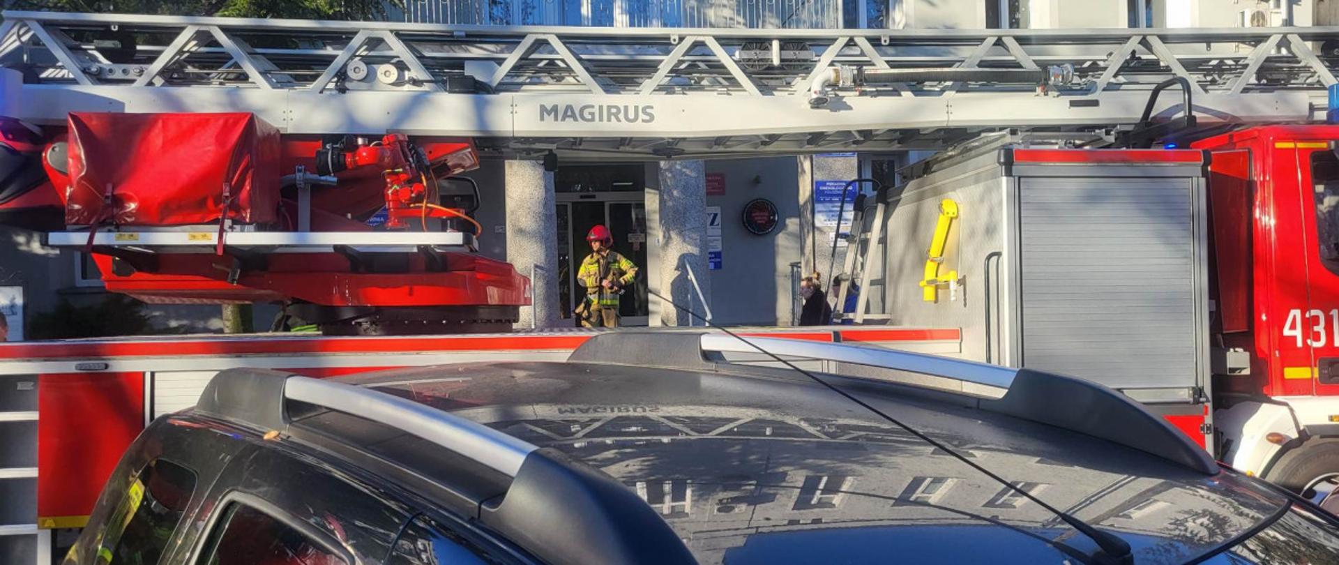 Drabina pożarnicza stoi przed szpitalem. Ze szpitala wychodzi strażak.