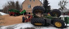 Widać przewrócony ciągnik, od przodu znajdują się koła, w pobliżu budynku jest przyczepa, na poboczu rozsypane zboże