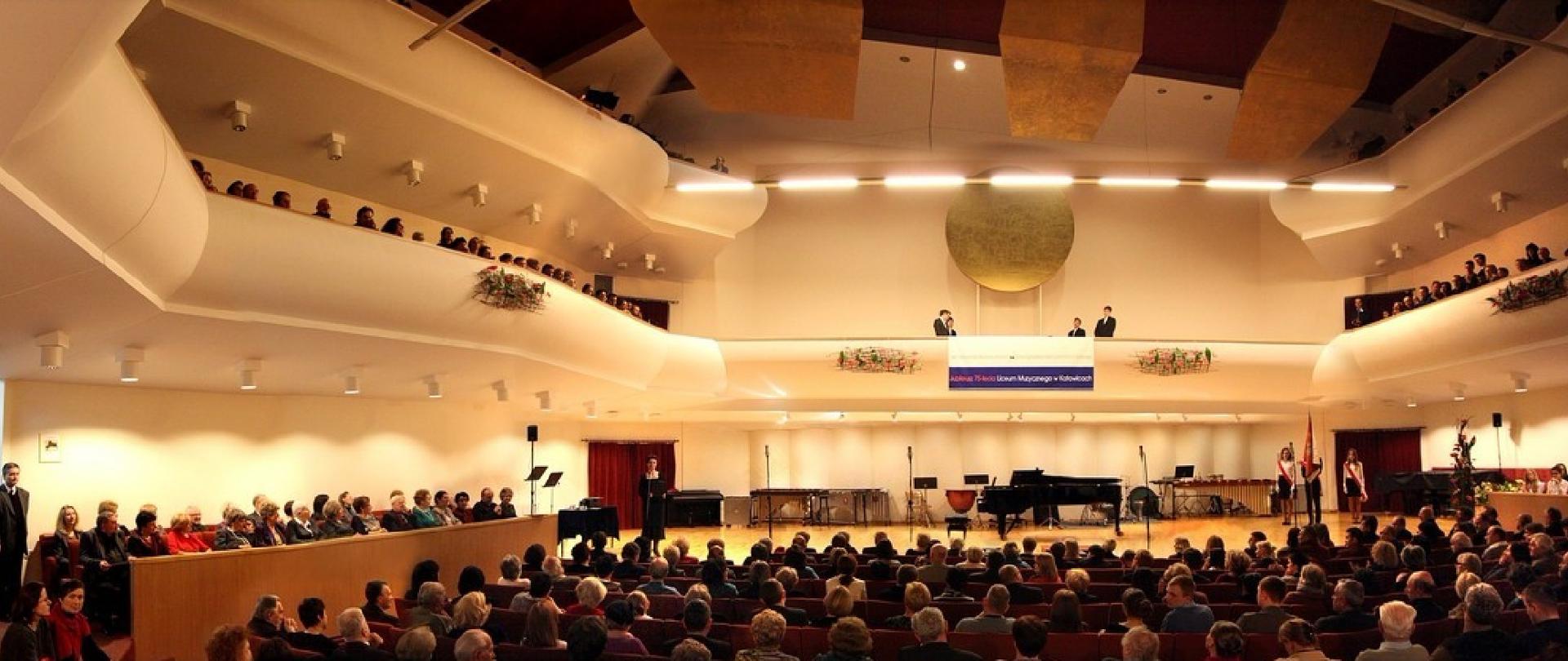 Sala koncertowa ZPSM - widok od strony publiczności, na scenie poczet sztandarowy oraz przemawiająca dyrektor ZPSM