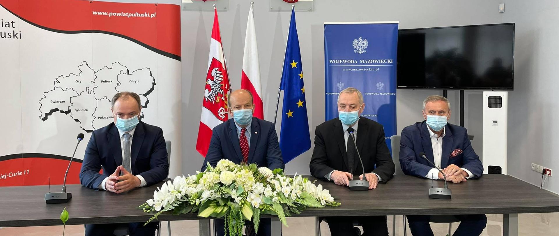 Na zdjęciu znajduje się czterech mężczyzn siedzących za stołem, przed sobą mają mikrofony, za nimi znajduje się między innymi flaga Polski i Unii Europejskiej. 