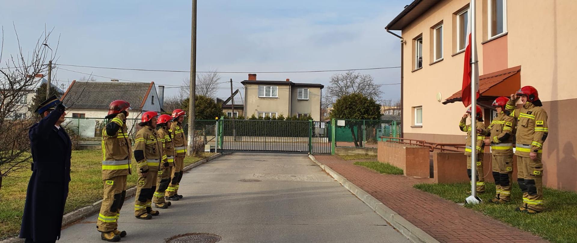 Przy maszcie flagowym stoi 3 strażaków w żółtych mundurach, jeden z nich podnosi flagę. Po drugiej stronie stoi 5 strażaków, 4 w żółtych mundurach jeden w granatowym płaszczu.