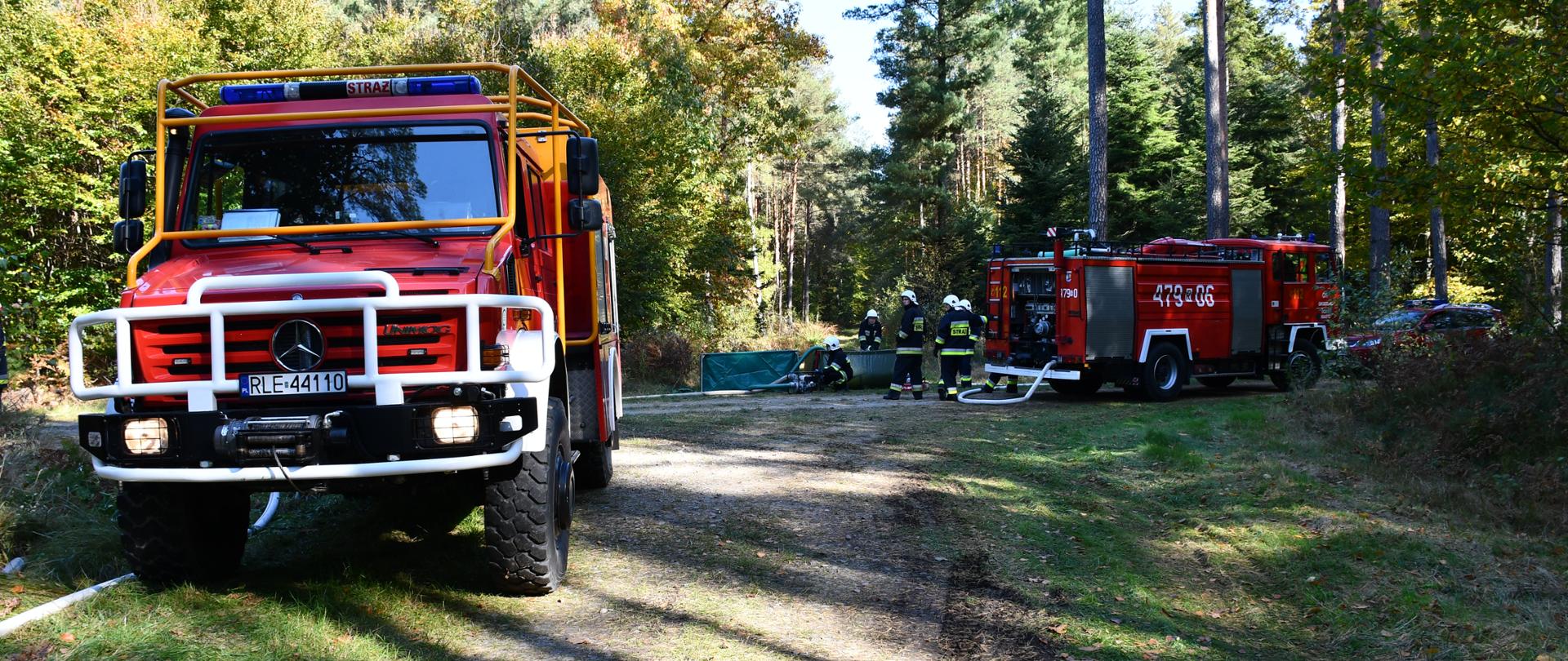Na zdjęciu widzimy samochody pożarnicze oraz strażaków rozkładających zbiornik na wodę. W tle widzimy drogę leśna i las.