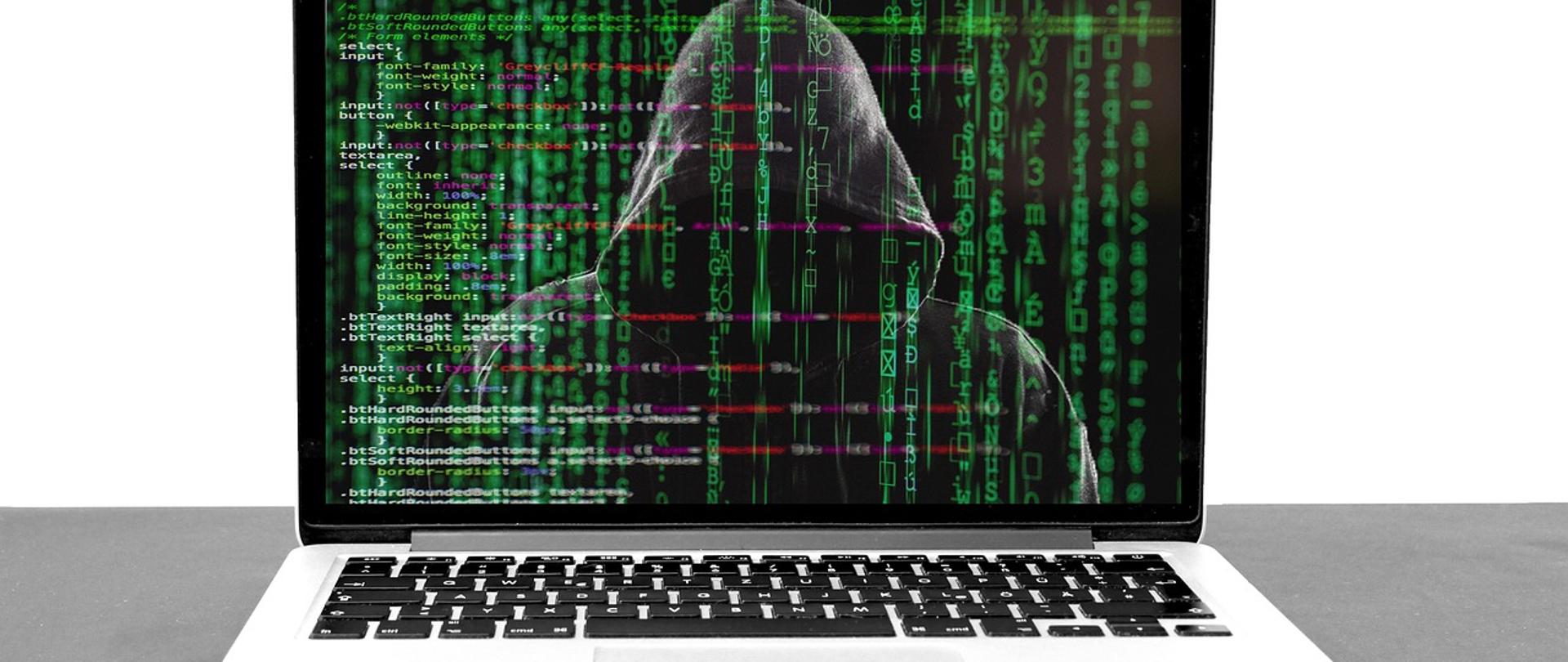 Zdjęcie laptopa. Na ekranie laptopa zamaskowana osoba. W tle kody systemowe.