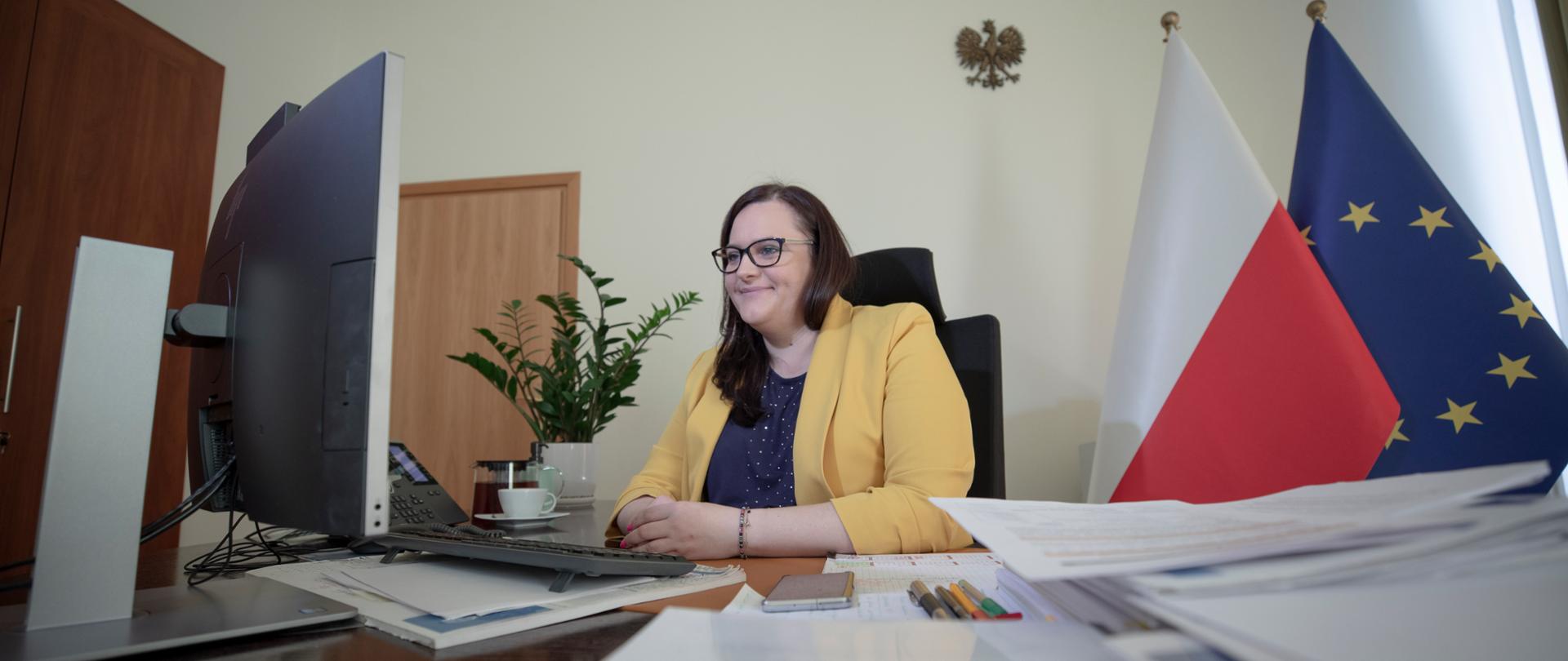 Wiceminister Małgorzata Jarosińska-Jedynak siedzi w swoim gabinecie przed monitorem. Na blacie biurka dokumenty. Z tyłu kwiat w doniczce i flagi PL i UE.