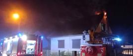 POŻAR PODDASZA. Na zdjęciu widać samochody strażackie stojące przy budynku, widać wydobywające się płomienie i dym z dachu, strażacy prowadzą działania ratowniczo-gaśnicze