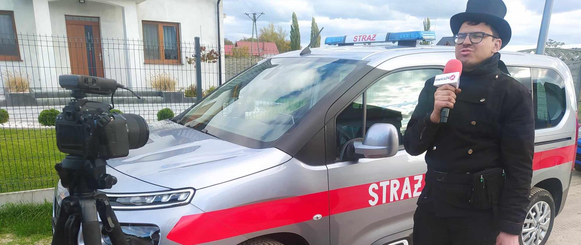 Czujka na straży Twojego bezpieczeństwa. Zdjęcie przedstawia kominiarza stojącego na tle samochodu strażackiego. Kominiarz ubrany w mundur kominiarski, mający na głowie czarny kapelusz, trzyma w prawej ręce mikrofon. Przed nim, na stojaku znajduje się kamera. Trwa nagrywanie spotu edukacyjnego. W tle widoczny jest fragment budynku mieszkalnego.