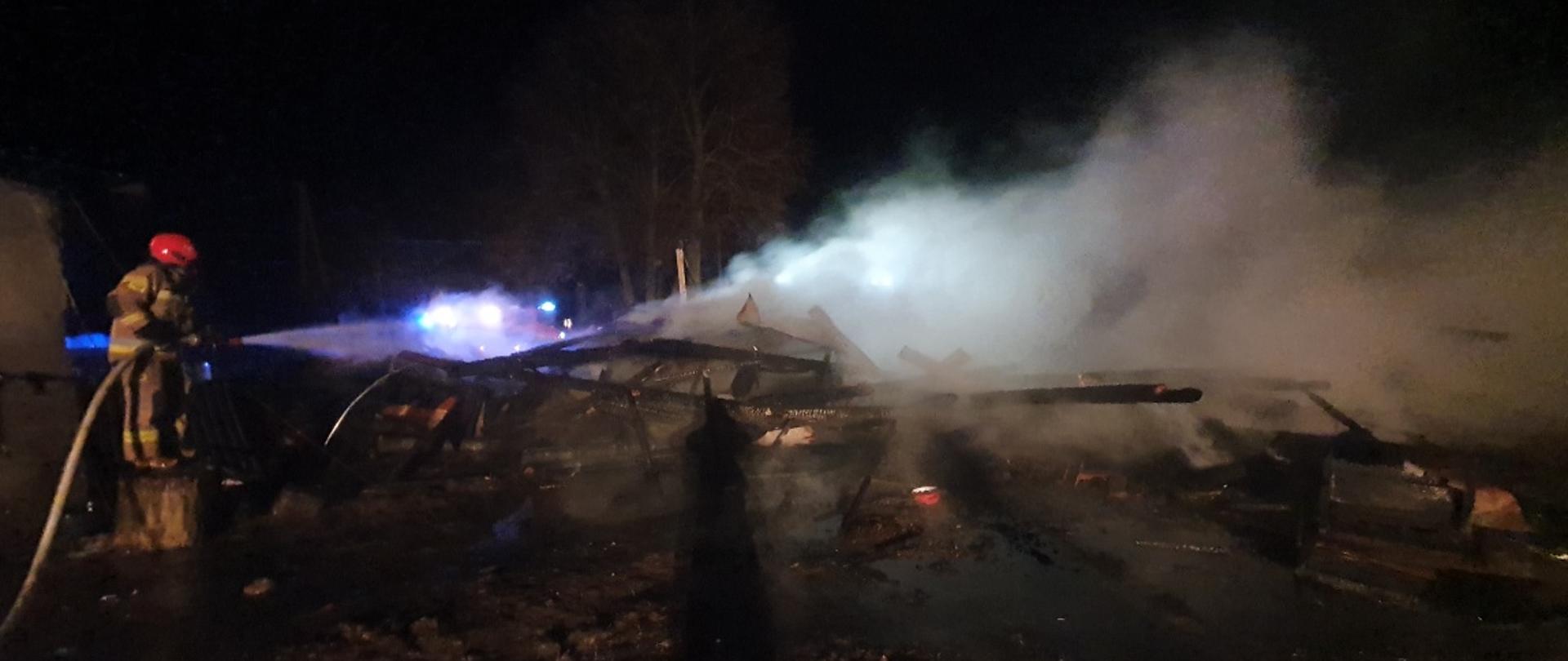 Strażak PSP z prądem gaśniczym polewa palące się zgliszcza. Dookoła panuje ciemność.
