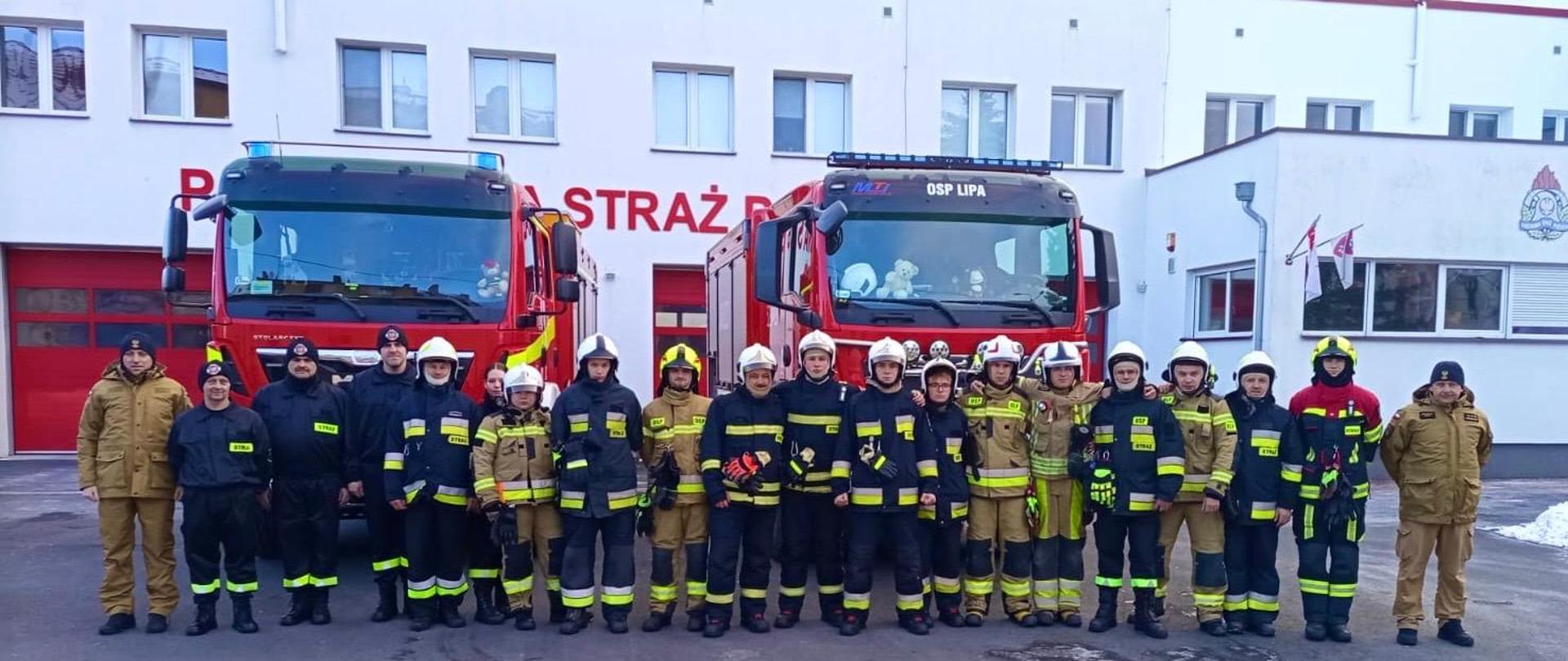 Grupa strażaków w umundurowaniu ochronnym stoi w szeregu przed remizą strażacką. Za nimi zaparkowane są dwa czerwone wozy strażackie, a na budynku widoczny jest napis „Straż”. Strażacy mają hełmy i odblaskowe elementy, pozują do wspólnego zdjęcia na zewnątrz.