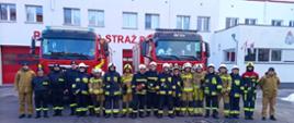 Grupa strażaków w umundurowaniu ochronnym stoi w szeregu przed remizą strażacką. Za nimi zaparkowane są dwa czerwone wozy strażackie, a na budynku widoczny jest napis „Straż”. Strażacy mają hełmy i odblaskowe elementy, pozują do wspólnego zdjęcia na zewnątrz.