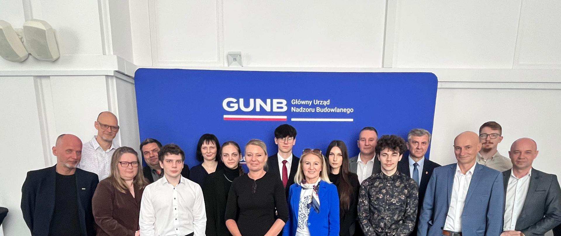 Grupowe zdjęcie uczestników spotkania z laureatami Ogólnopolskiej Olimpiady Wiedzy o Procesie Inwestycyjno-Budowlanym. Uczniowie, opiekunowie i przedstawiciele GUNB stoją na tle ścianki z logo Głównego Urzędu Nadzoru Budowlanego