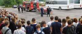 Na zdjęciu funkcjonariusz Policji i Straży pożarnej , w tle widać samochód strażacki z podczepioną łodzią ratowniczą. W okół Strażaka i Policjanta zgromadzone dzieci szkoły podstawowej.