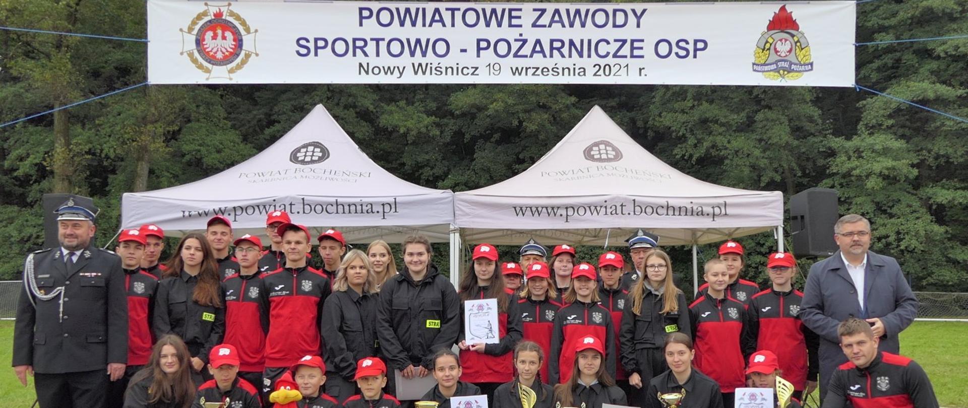Zdjęcie w centralnej części przedstawia grupę strażaków z Ochotniczej Straży Pożarnej w Mikluszowicach. Po lewej stronie zdjęcia stoi strażak ubrany w strój galowy. Po prawej stronie zdjęcia znajduje się Starosta Powiatu Bocheńskiego. W tle widać baner „Powiatowe Zawody Sportowo Pożarnicze OSP”