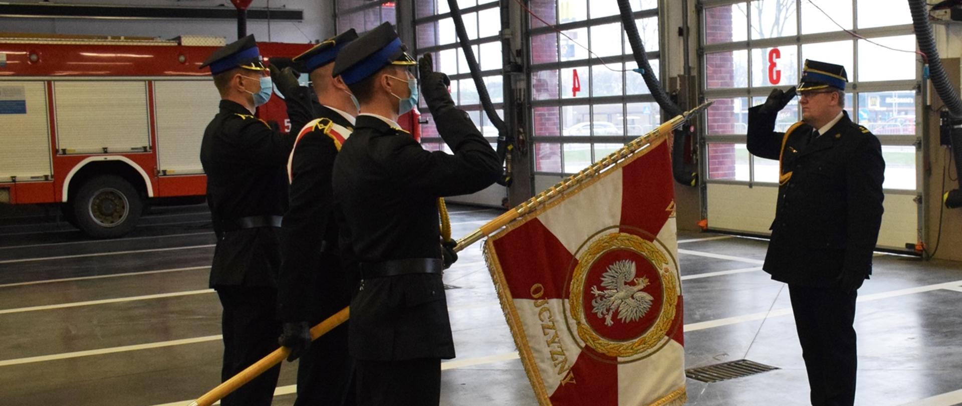 Oficer Państwowej Straży Pożarnej, w ubraniu wyjściowym salutuje przed sztandarem. Sztandar w rękach pocztu sztandarowego złożonego z trzech strażaków. 