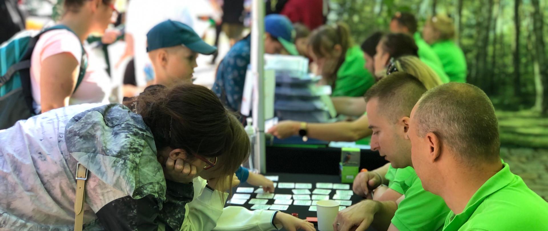 Na stołach pomoce edukacyjne, z jednej strony osoby w zielonych koszulkach, z drugiej dzieci i dorośli, w tle ściana namiotu 