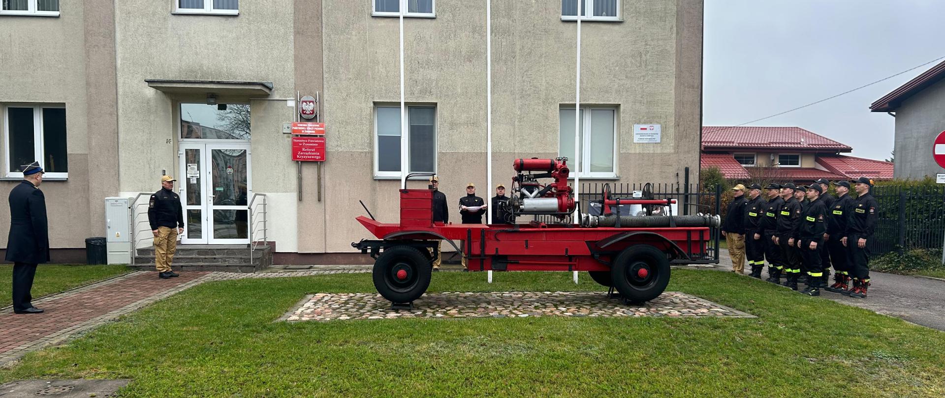 Zdjęcie ukazuje uroczystość z okazji Dnia Niepodległości przed budynkiem Komendy Powiatowej Państwowej Straży Pożarnej w Żurominie, z widocznym historycznym sprzętem gaśniczym na centralnym planie. Po prawej stronie w szeregu stoi grupa strażaków w czarnych mundurach, a po lewej dwóch dowódców lub honorowych gości, z których jeden jest w galowym mundurze, co podkreśla uroczysty charakter wydarzenia.