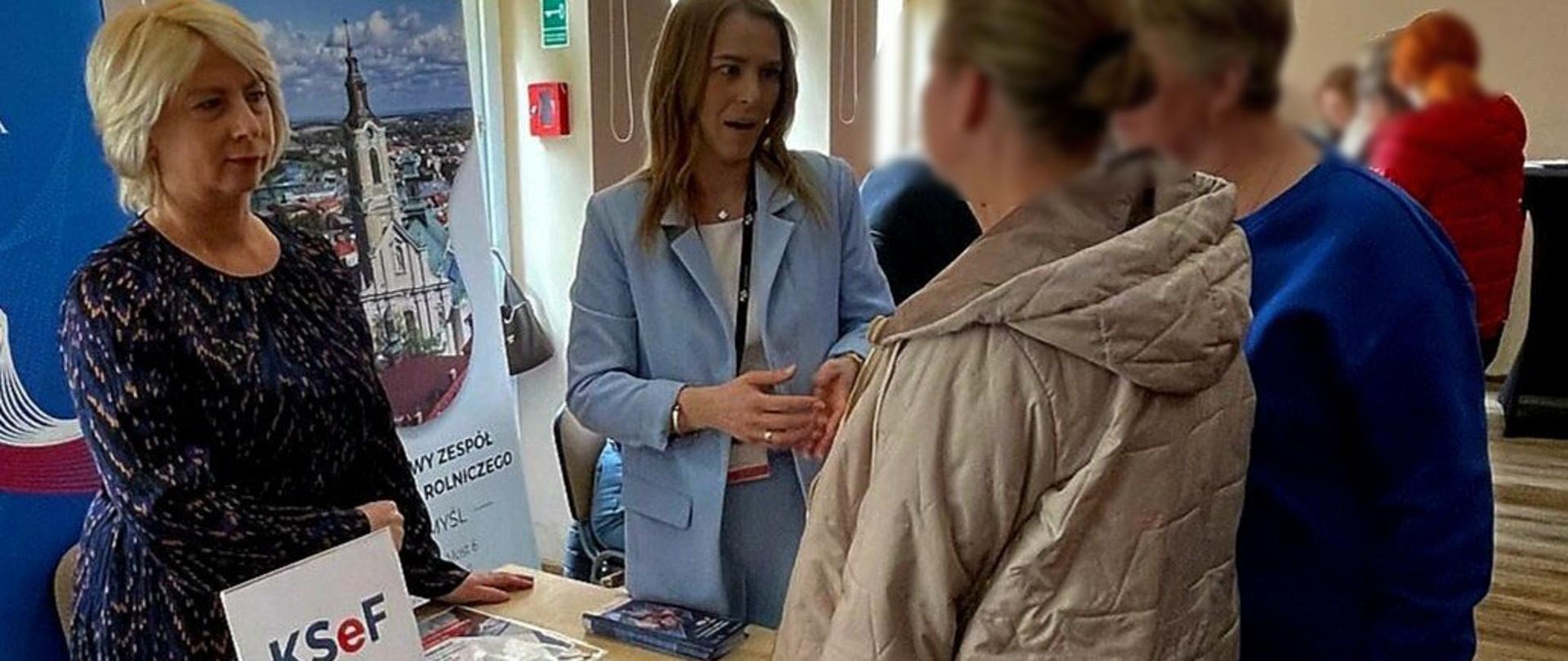 Ekspertki KAS stoją przy stanowisku informacyjnym. Przed nimi są uczestnicy konferencji. Na stoliku leżą ulotki. 