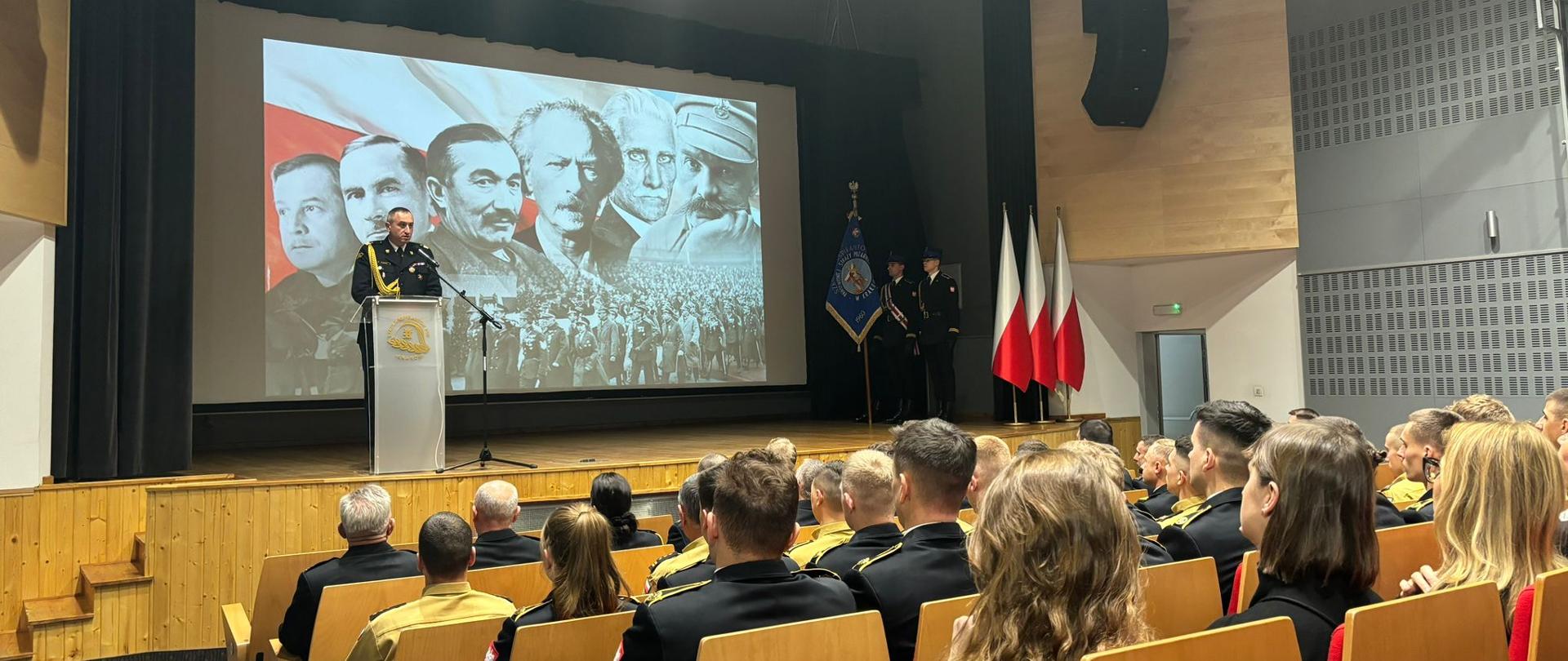 3. Zdjęcie wykonane na auli Szkoły Aspirantów PSP w Krakowie. W tle na białym ekranie wyświetla się zdjęcie które przedstawia sławnych Polaków zasłużonych podczas wydarzeń niepodległościowych. Na zdjęciu są widoczni funkcjonariusze którzy siedzą tyłem do fotografa na krzesłach w auli. Na scenie komendant szkoły w granatowym mundurze mówi do zgromadzonych. 