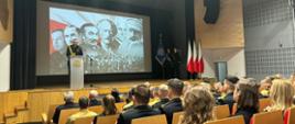 Zdjęcie wykonane na auli Szkoły Aspirantów PSP w Krakowie. W tle na białym ekranie wyświetla się zdjęcie które przedstawia sławnych Polaków zasłużonych podczas wydarzeń niepodległościowych. Na zdjęciu są widoczni funkcjonariusze którzy siedzą tyłem do fotografa na krzesłach w auli. Na scenie komendant szkoły w granatowym mundurze mówi do zgromadzonych. 