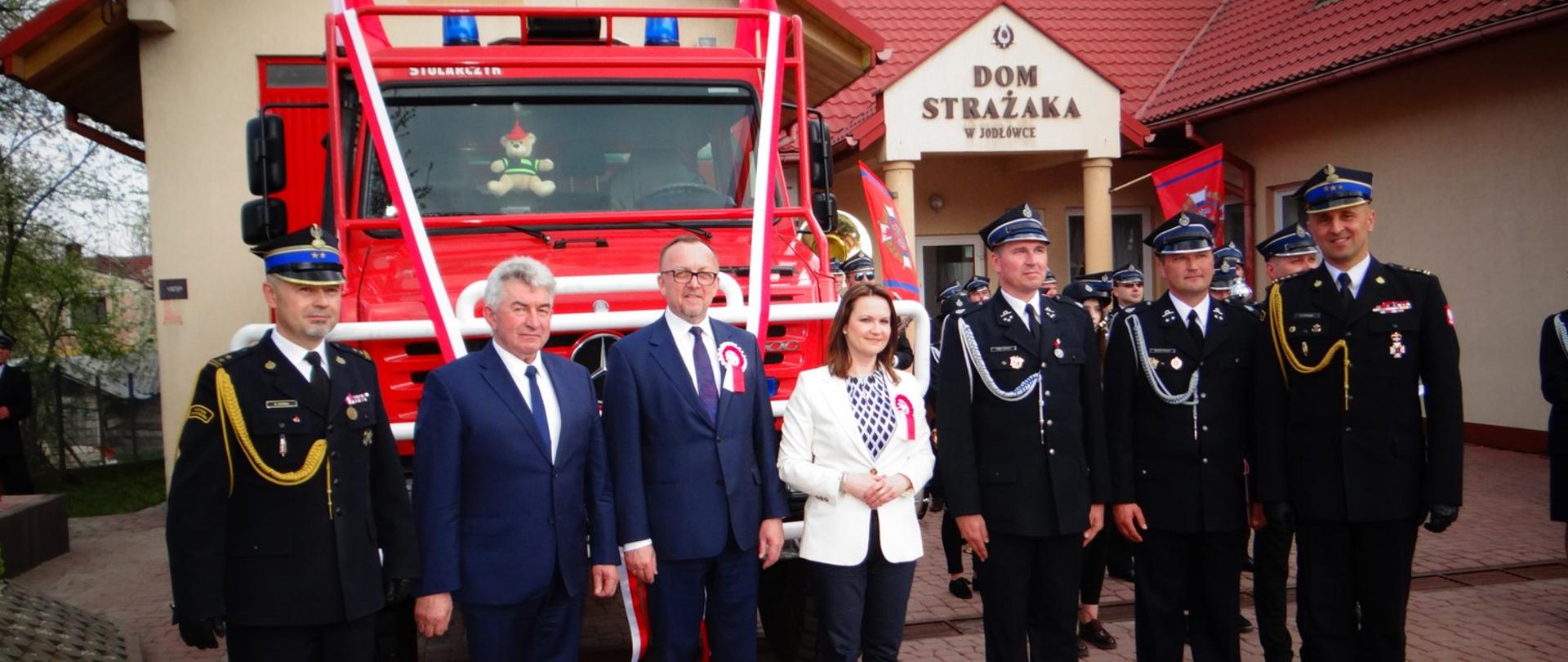 Na zdjęciu widzimy przedstawicieli władz rządowych i samorządowych, zastępcę podkarpackiego komendanta wojewódzkiego PSP oraz komendanta powiatowego PSP stojących przed przekazanym samochodem pożarniczym Mercedes Unimog. W tle widać strażaków OSP oraz remizę OSP Jodłówka.