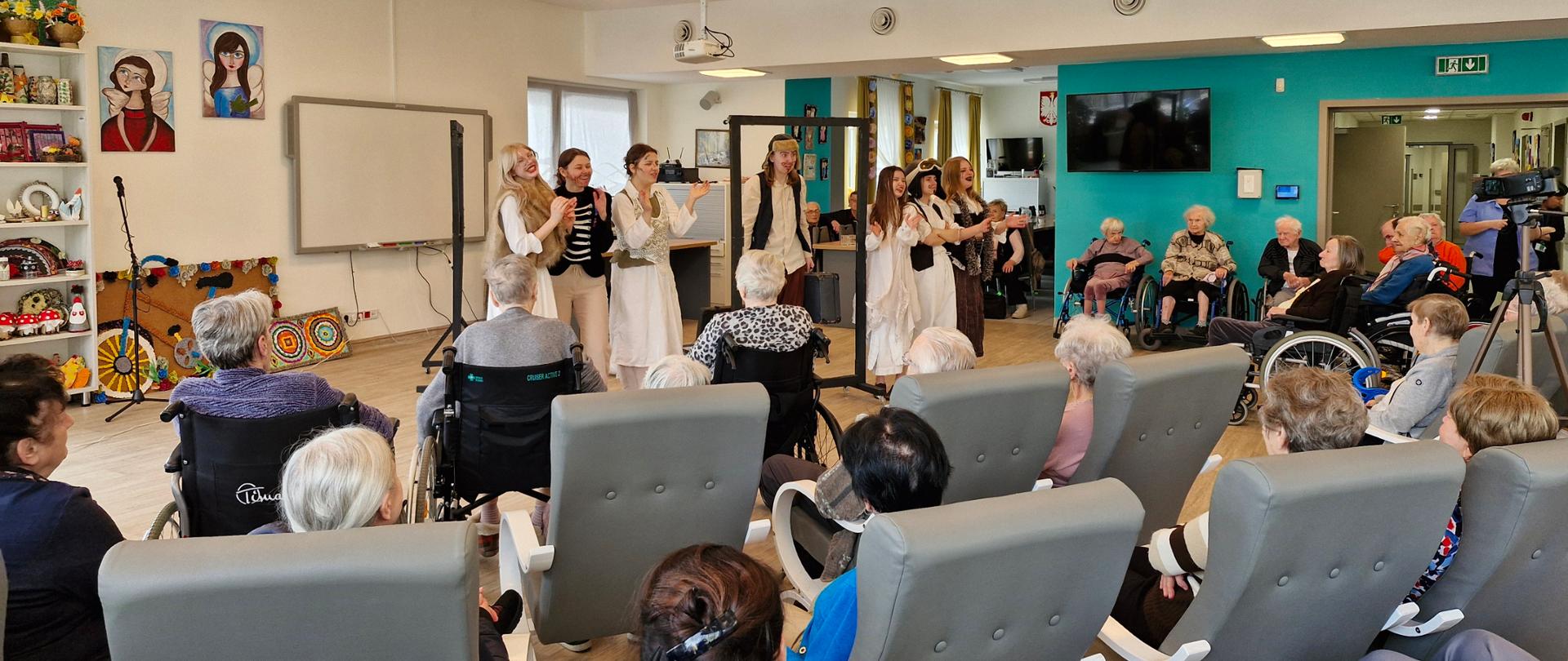 W dużej jasnej sali stoi i klaszcze siedem dziewcząt w biało - czarno-beżowych kostiumach teatralnych i klaszczą. Z przodu i z boku widać osoby, głównie starsze siedzące na fotelach lub wózkach inwalidzkich. Po prawej stronie stoi kamera na statywie. Na ścianach widać kolorowe obrazy, regał z kolorowymi ozdobami, telewizory, okna.