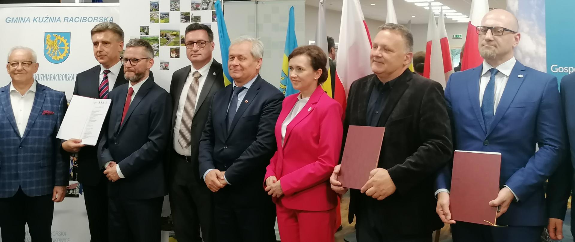 Podpisanie listu intencyjnego ws. budowy Zbiornika Kotlarnia. Na zdjęciu 8 osób, stojących w rzędzie koło siebie. Wśród nich jedna kobieta, ubrana w różową garsonkę. W tle roll upy z nazwami partnerów projektu.