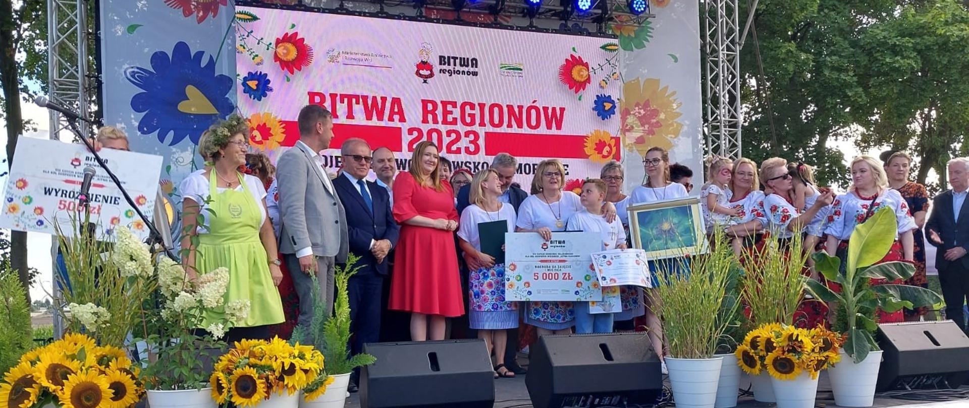Laureatki i laureaci konkursu, na scenie z symbolicznymi czekami i dyplomami. Dwie kobiety z dziećmi na rękach.