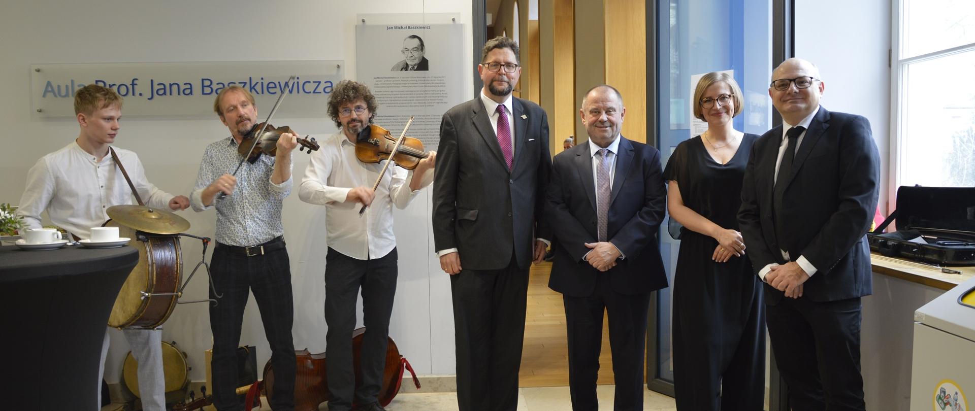 W sali stoi w rzędzie grupa czterech osób, pośrodku wiceminister Szeptycki, z boku trzech mężczyzn w białych koszulach gra na instrumentach.