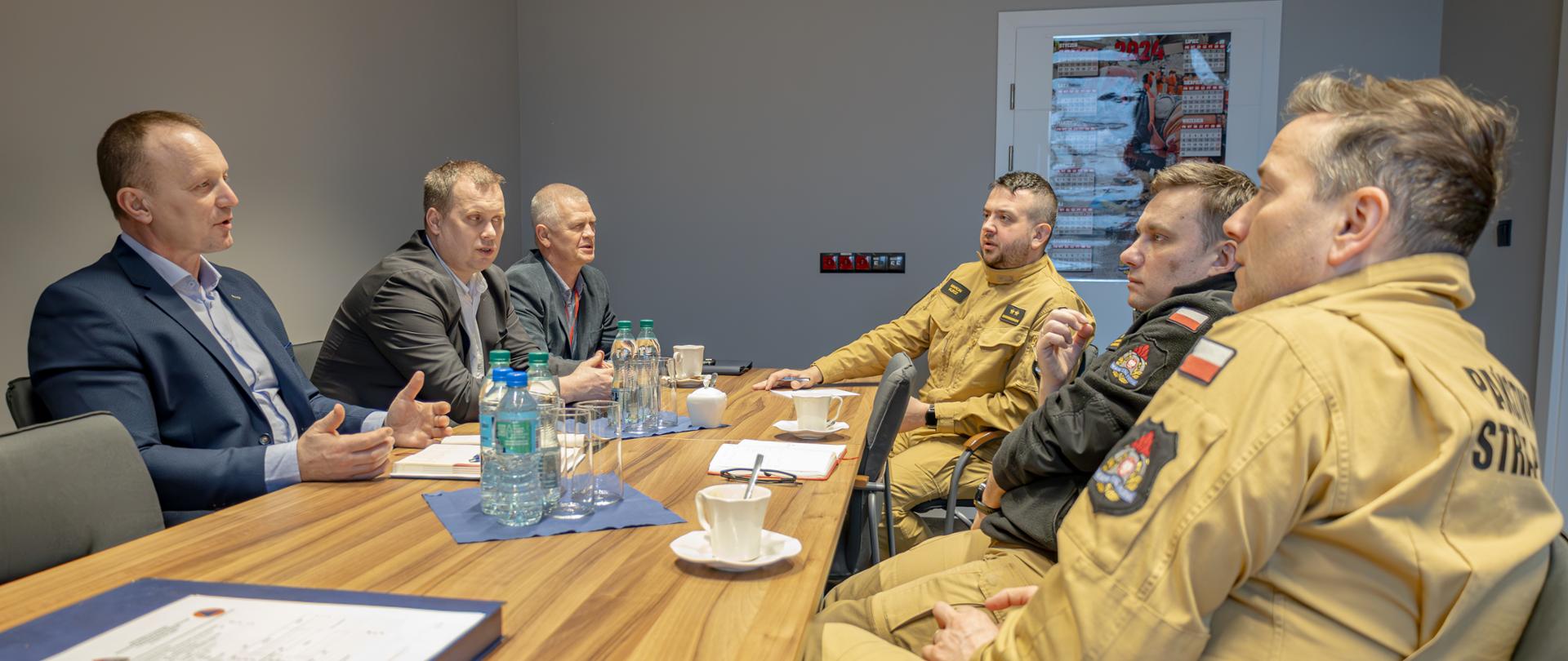 Mężczyźni w garniturach i strażacy siedzą przy stole konferencyjnym, prowadzą ze sobą dyskusję.