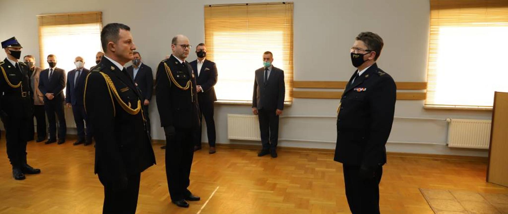 Zdjęcie przedstawia Zastępcę Podlaskiego Komendanta Wojewódzkiego Państwowej Straży Pożarnej starszego brygadiera Roberta Dzierżka podczas przyjmowania meldunku od zdającego obowiązki Komendanta Powiatowego Państwowej Straży Pożarnej w Zambrowie brygadiera Macieja Krajewskiego oraz nowo powołanego Komendanta Powiatowego Państwowej Straży Pożarnej w Zambrowie starszego brygadiera Krzysztofa Konickiego. Komendanci ubrani w mundury wyjściowe ze sznurem galowym. Uroczystość odbywała się w jednym z pomieszczeń Komendy Powiatowej Państwowej Straży Pożarnej w Zambrowie.