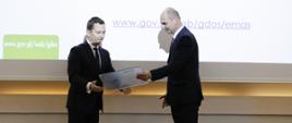 Widać dwóch mężczyzn ubranych w garnitury, białe koszule i krawaty. Jednym z nich jest Generalny Dyrektor Ochrony Środowiska - Andrzej Szweda - Lewandowski, który wręcza certyfikat EMAS.