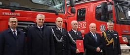 Strażacy ochotniczej straży pożarnej oraz Państwowej Straży Pożarnej stoją obok siebie za nimi stoją dwa samochody gaśnicze. Strażak ochotniczej straży pożarnej trzyma czerwoną teczkę.