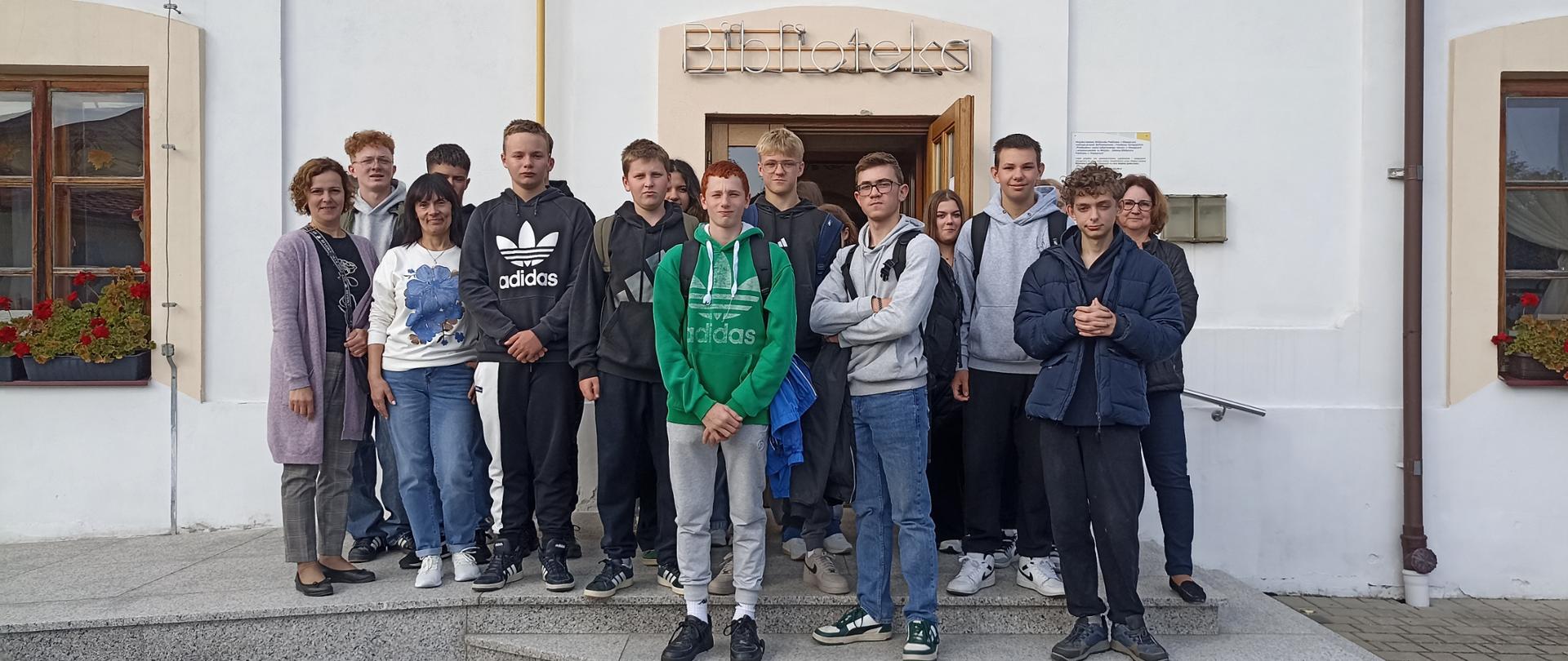 Grupa kilkunastu młodych osób i dwóch dorosłych stoi na schodach przed wejściem do budynku z napisem „Biblioteka”. Uczestnicy są ubrani na co dzień, część ma na sobie bluzy z kapturem. W tle widać jasną elewację budynku, okna z kwiatami oraz drzwi wejściowe.