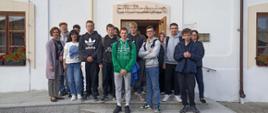 Grupa kilkunastu młodych osób i dwóch dorosłych stoi na schodach przed wejściem do budynku z napisem „Biblioteka”. Uczestnicy są ubrani na co dzień, część ma na sobie bluzy z kapturem. W tle widać jasną elewację budynku, okna z kwiatami oraz drzwi wejściowe.
