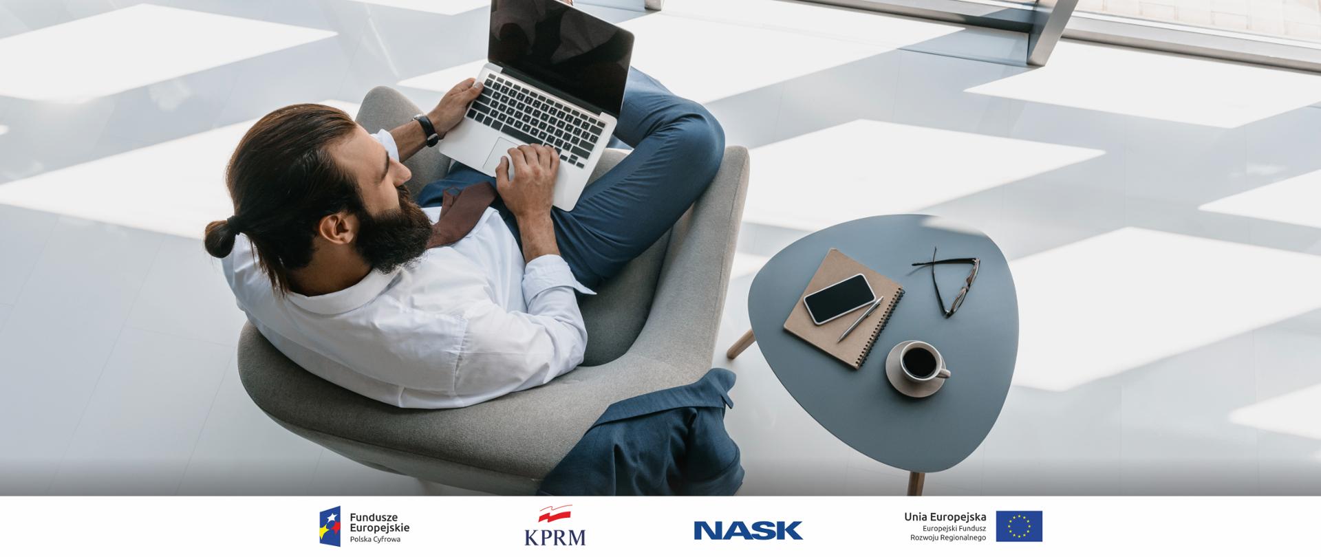 Zdjęcie, ujęcie od góry - mężczyzna w jasnym pokoju, siedzi w fotelu, korzysta z laptopa, obok fotela stolik, na którym stoi filiżanka z kawą, leżą zeszyt, smartfon i okulary.