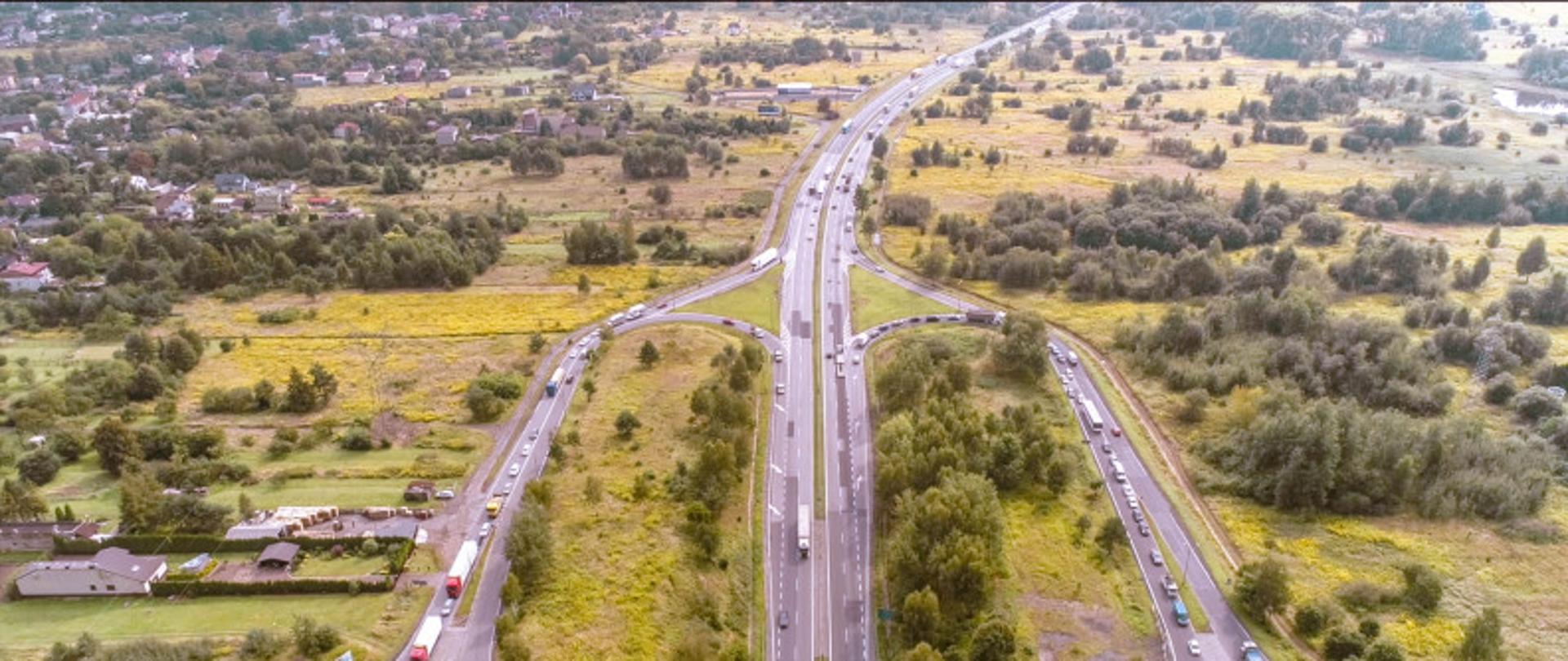 Zdjęcie przedstawia widok z lotu drona na węzeł drogowy na drodze ekspresowej S1. Widoczne poruszające się samochody, w tle mała miejscowość i zadrzewienia.