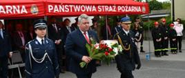 Na zdjęciu widzimy uroczystość z udziałem przedstawicieli służb mundurowych, odbywającą się przed budynkiem oznaczonym czerwonym namiotem z napisem „PAŃSTWOWA STRAŻ POŻARNA”. Na pierwszym planie idzie trójka osób: po lewej stronie kobieta w granatowym mundurze policyjnym, w środku mężczyzna w garniturze z przypiętą biało-czerwoną kokardą narodową, trzymający w rękach wiązankę biało-czerwonych kwiatów z liśćmi palmowymi, po prawej stronie mężczyzna w galowym mundurze strażackim z ozdobnymi sznurami i czapką. W tle pod namiotem stoi grupa oficjeli i gości w garniturach, a po prawej stronie widoczna jest grupa młodzieży w mundurach Młodzieżowych Drużyn Pożarniczych z czerwonymi czapkami oraz kobieta trzymająca dziecko. Uroczystość ma charakter patriotyczny, strażacki. 