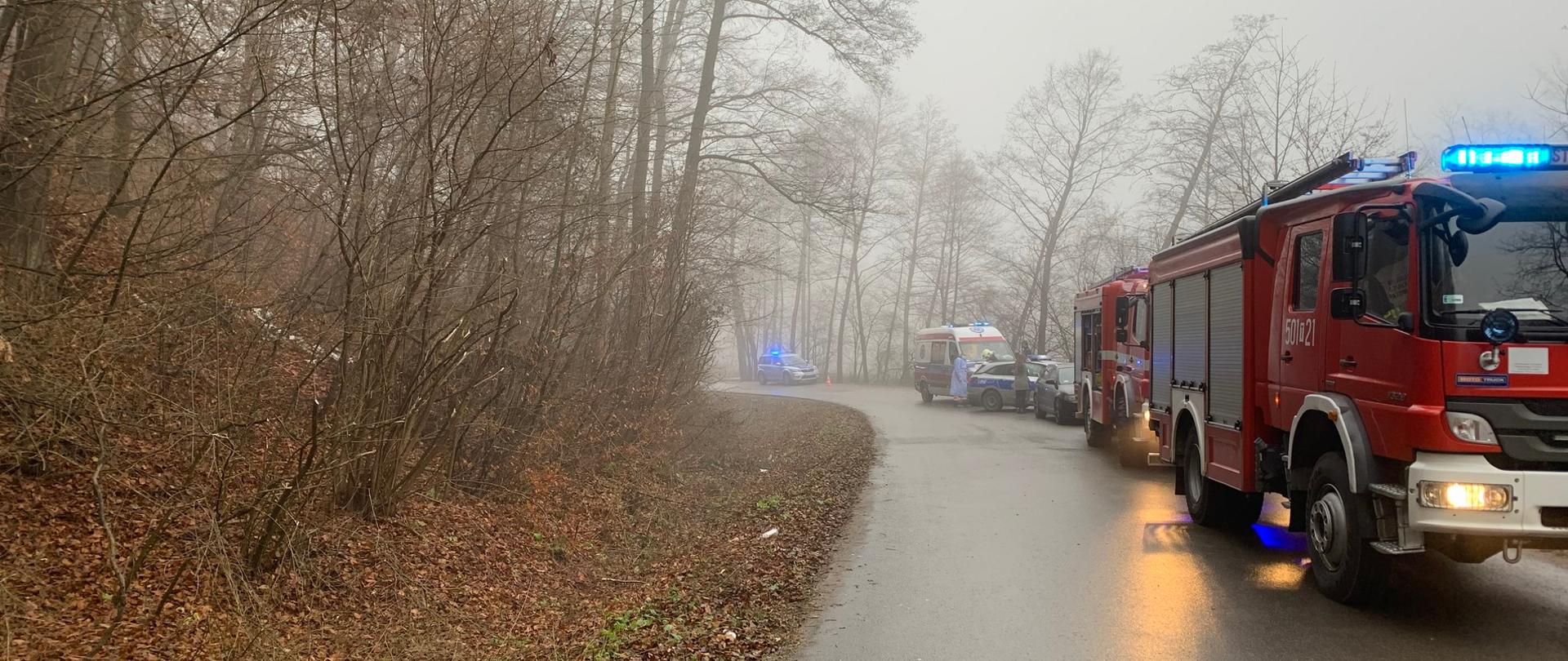 Na łuku drogi ustawione są na jednym pasie pojazdy służb ratowniczych. Na pierwszym planie widać pojazd pożarniczy z włączoną sygnalizacja świetlną.