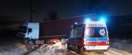 Samochód ciężarowy po wypadku. Ciągnik wjechał do rowu. Naczepa stoi w poprzek jezdni. Przed naczepą stoi ambulans pogotowia ratunkowego z włączonymi światłami uprzywilejowania.