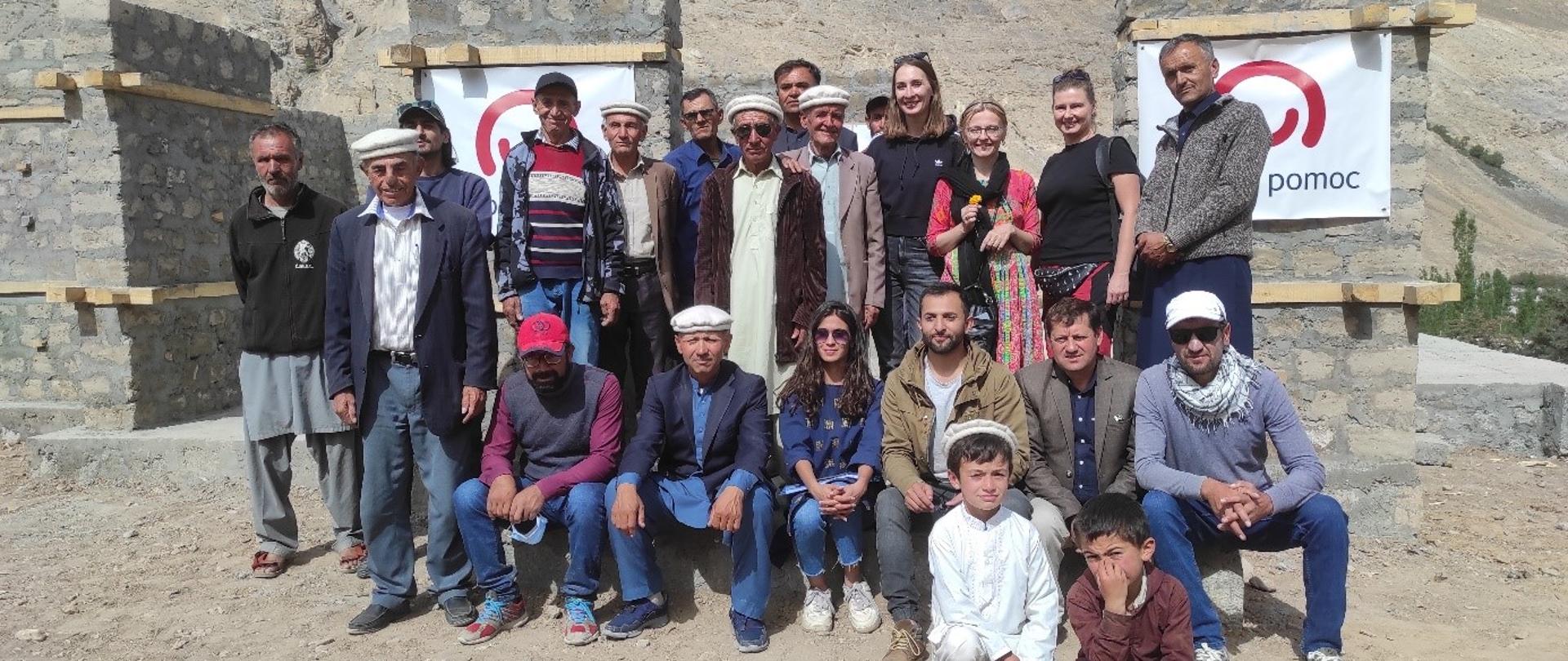 Grupa ludzi pozuje przy fundamentach budynku na tle gór. Na fundamentach widnieje logo z napisem polska pomoc.