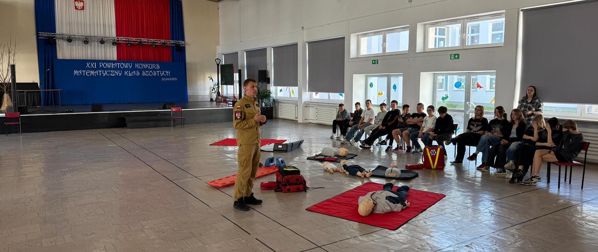 Na zdjęciu widać strażaka w mundurze prowadzącego pokaz udzielania pierwszej pomocy w sali gimnastycznej lub auli szkolnej. Przed nim na matach ułożone są fantomy treningowe oraz sprzęt ratowniczy – torby medyczne, deska ortopedyczna i szyny. Naprzeciwko strażaka siedzi kilkunastoosobowa grupa młodzieży, uważnie słuchająca i obserwująca przebieg zajęć. 