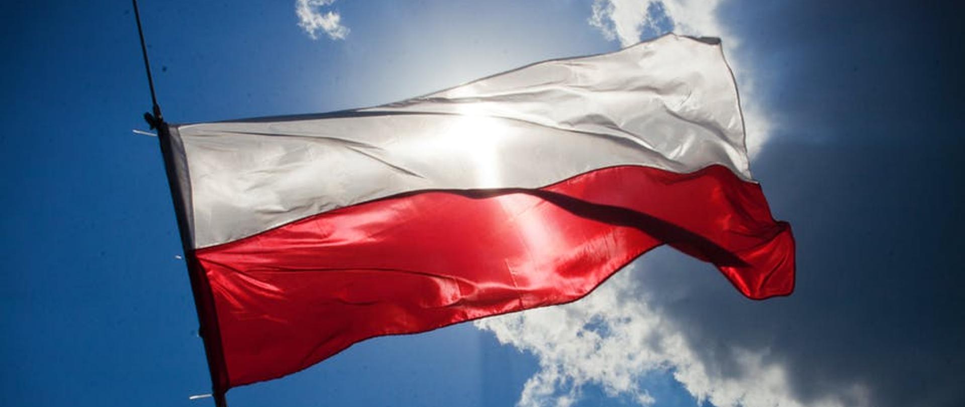 Flaga Polski na tle nieba z chmurami, przez którą przebijają promienie słońca.