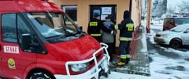 Kolorowe zdjęcie przedstawia na pierwszym planie, po lewej, część przednią czerwonego samochodu lekkiego Ochotniczej Straży Pożarnej z napisem Straż. Na środkowym planie widoczne dwie postacie umundurowanych na czarno strażaków z napisami na plecach Straż, podtrzymujących rękami za plecy starsza osobę pośrodku nich, pomagających wejść do budynku przez drzwi z napisem Punkt Szczepień. Na zdjęciu widoczny w wielu miejscach śnieg.