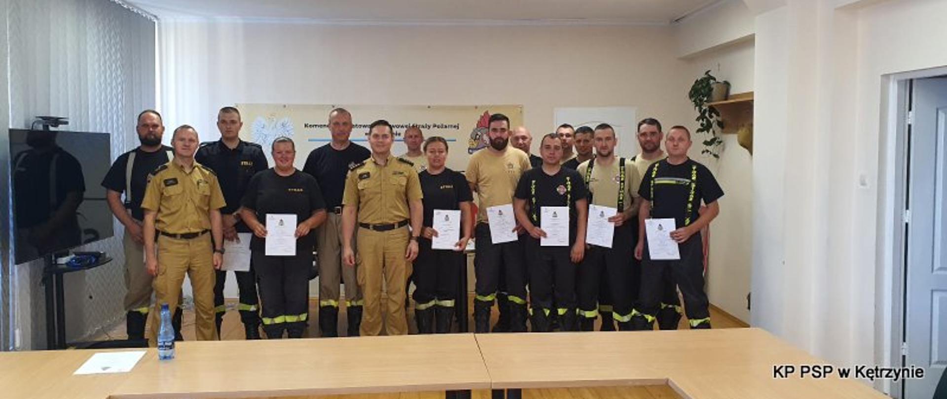Zdjęcie wykonane w Sali konferencyjnej kętrzyńskiej komendy. Strażacy ustawieni do zdjęcia po zdanym egzaminie. Kilkoro z nich trzyma przed sobą zaświadczenie z kursu.