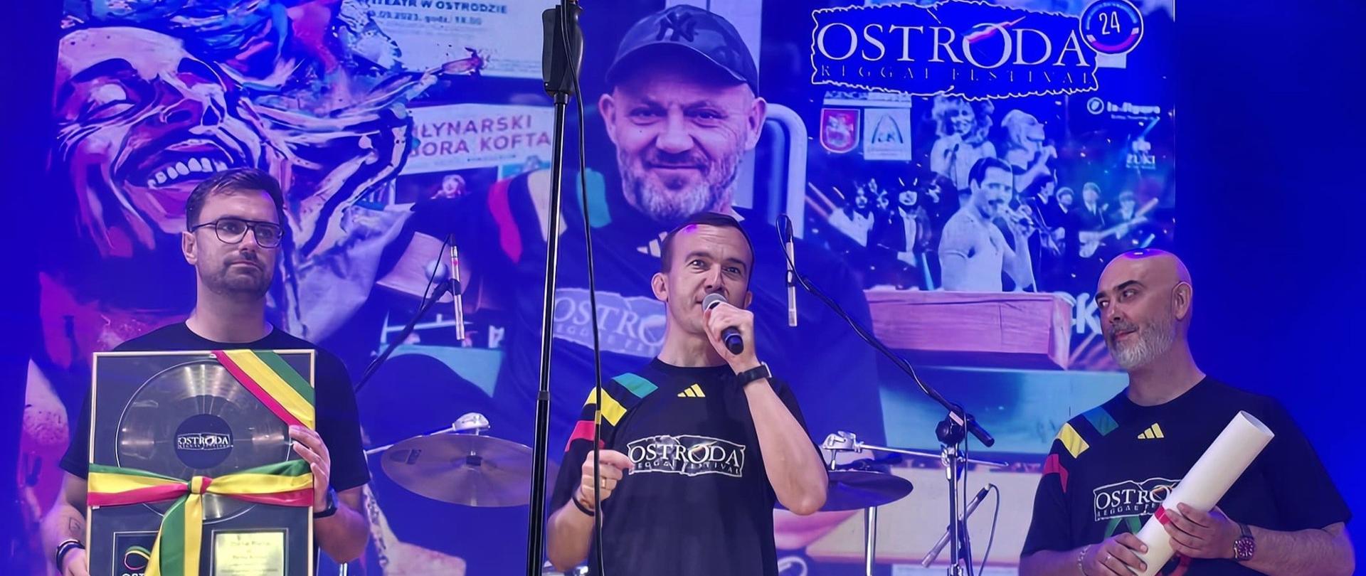 Trzech mężczyzn ubranych w czarne koszulki stoi na scenie, jeden z nich przemawia przez mikrofon 