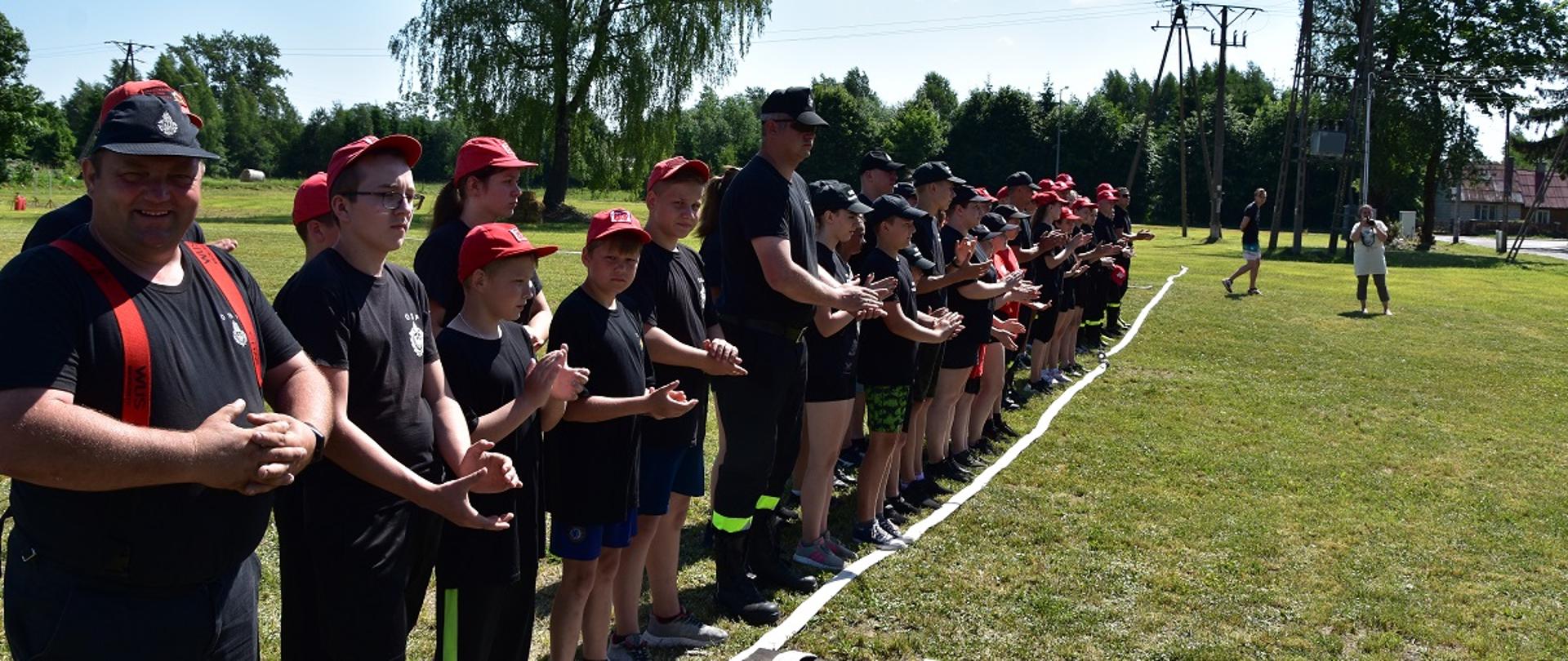 Zdjęcie obrazuje strażaków i młodzież ustawionych w dwuszeregu