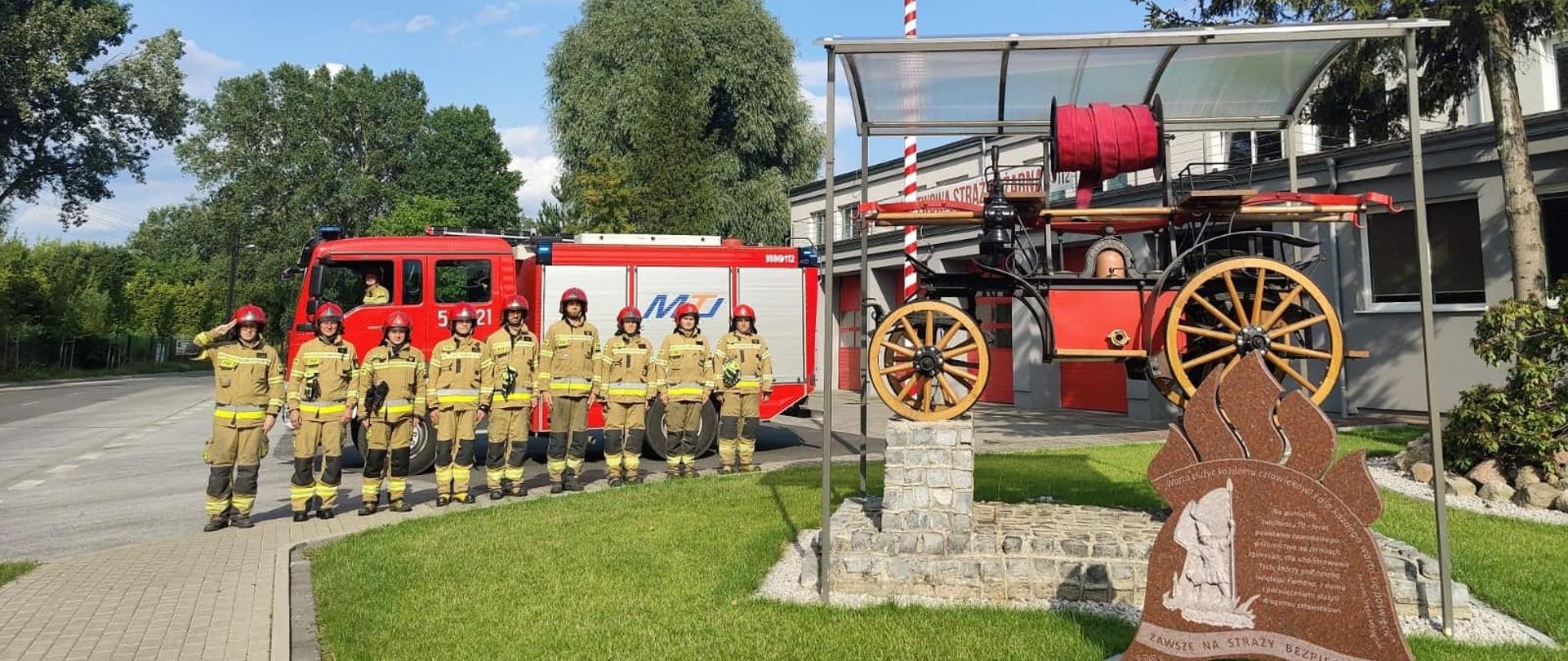 dziewięciu strażaków w ubraniach bojowych stojących na zbiórce przed garażem jednostki ratowniczo-gaśniczej za strażakami stoi samochód gaśniczy, przed strażakami z boku stoi zabytkowa sikawka oraz masz z flaga Polski 