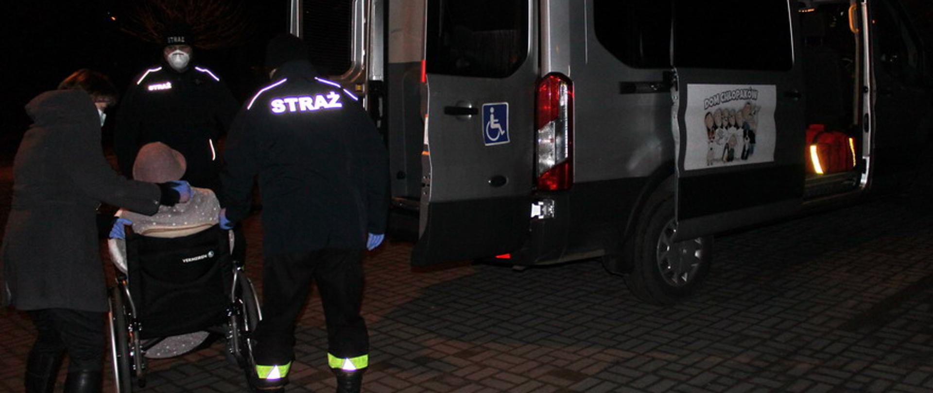 Dwóch strażaków wraz z opiekunem pomagają w transporcie na szczepienie starszej kobiecie poruszającej się na wózku inwalidzkim.