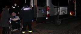 Dwóch strażaków wraz z opiekunem pomagają w transporcie na szczepienie starszej kobiecie poruszającej się na wózku inwalidzkim.