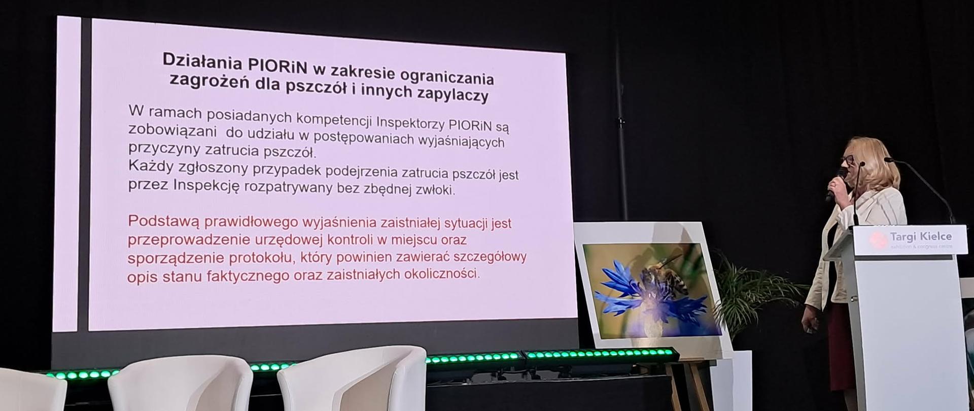 Inspektor na scenie omawia działania inspekcji w zakresie ograniczania zagrożeń dla pszczół i innych zapylaczy. W tle znajduje się prezentacja i zdjęcie pszczoły.